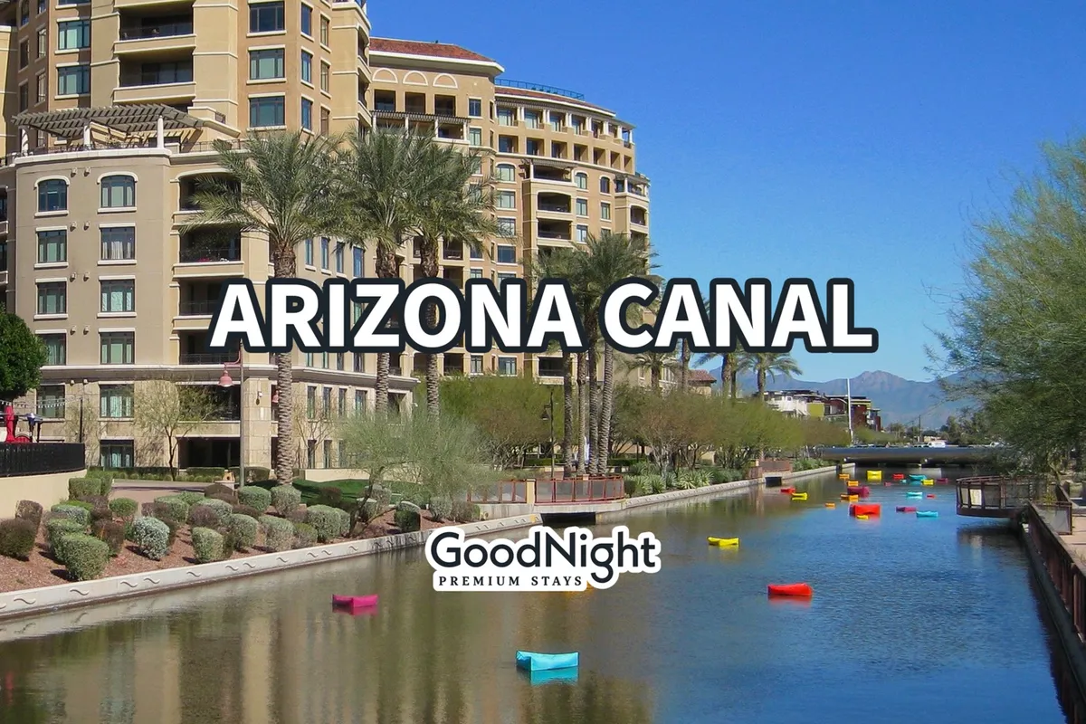 This image captures the Arizona Canal with colorful boats docked alongside the water, palm trees lining the banks, and nearby buildings that hint at vibrant community life. The cheerful atmosphere is highlighted by people strolling and the lively, waterfront ambiance.