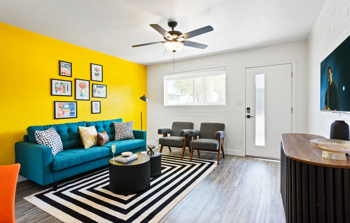 A bright, modern living room featuring a vibrant yellow accent wall, a plush teal sofa, a striped rug, a round coffee table, and contemporary artwork. The space is styled with pops of color and designed for socializing, reading, and relaxing, with a layout that encourages gathering.