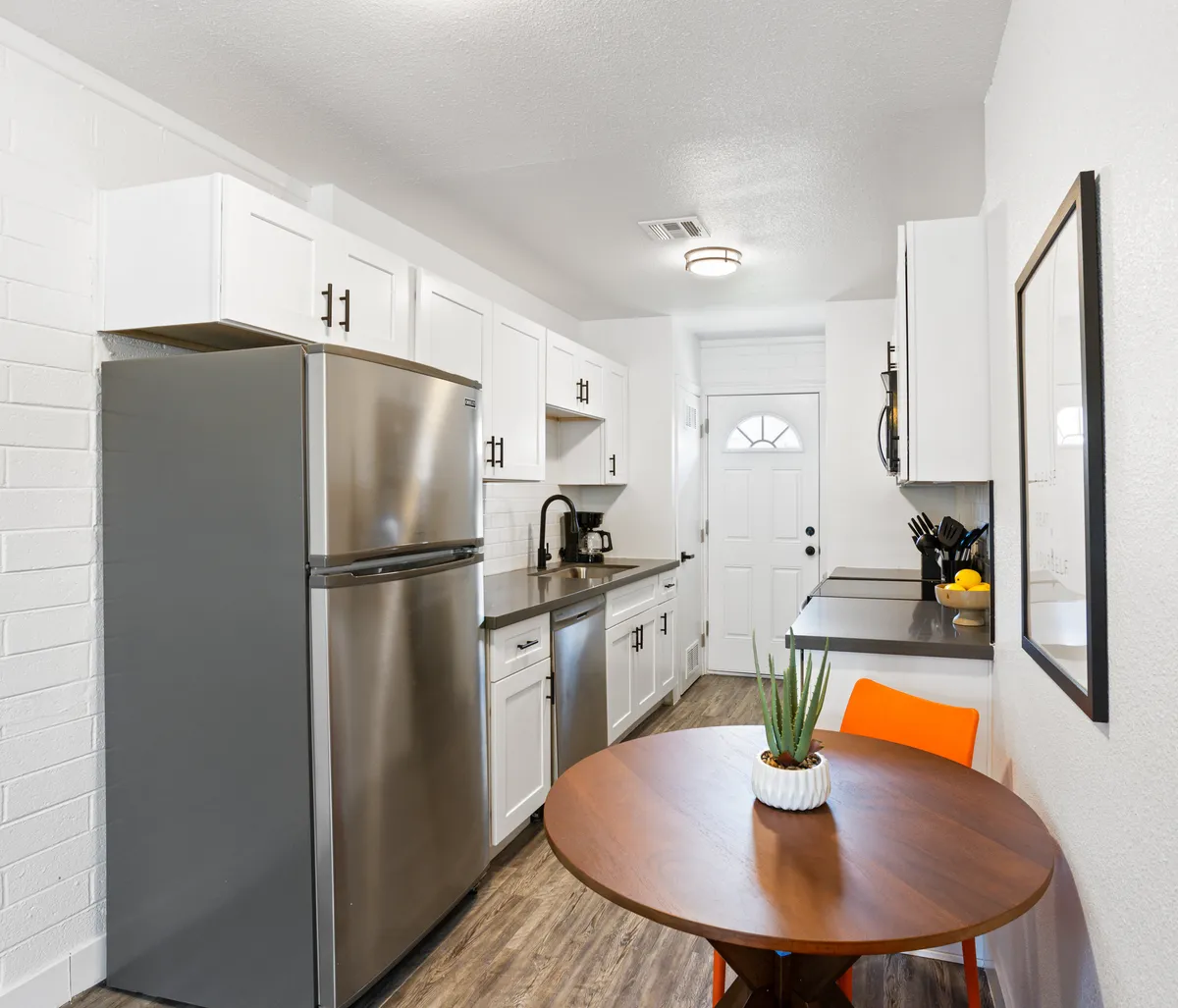 A cozy kitchen and dining nook scene with a stainless steel refrigerator, round wooden dining table, a pop of color from an orange chair, and a green plant for freshness. The layout invites intimate dinners or casual gatherings, emphasizing the connection between kitchen and dining area.