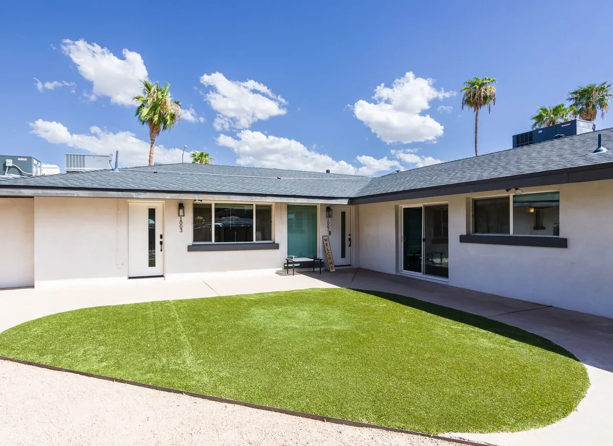 This image displays the inviting exterior of one of the homes, featuring a lush green lawn, tall palm trees, a clear blue sky, and the entrance door. The scene radiates relaxation and the ease of stepping into a welcoming residential space, while also connecting directly to the urban environment.