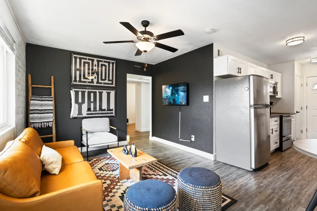 The image showcases a beautifully styled open-concept living area with a sleek leather sofa, vibrant blue poufs, elegant contemporary artwork, a central coffee table, and a modern kitchen in the background. The space exudes warmth and sophistication, perfect for gathering, relaxing, or celebrating with friends and family.