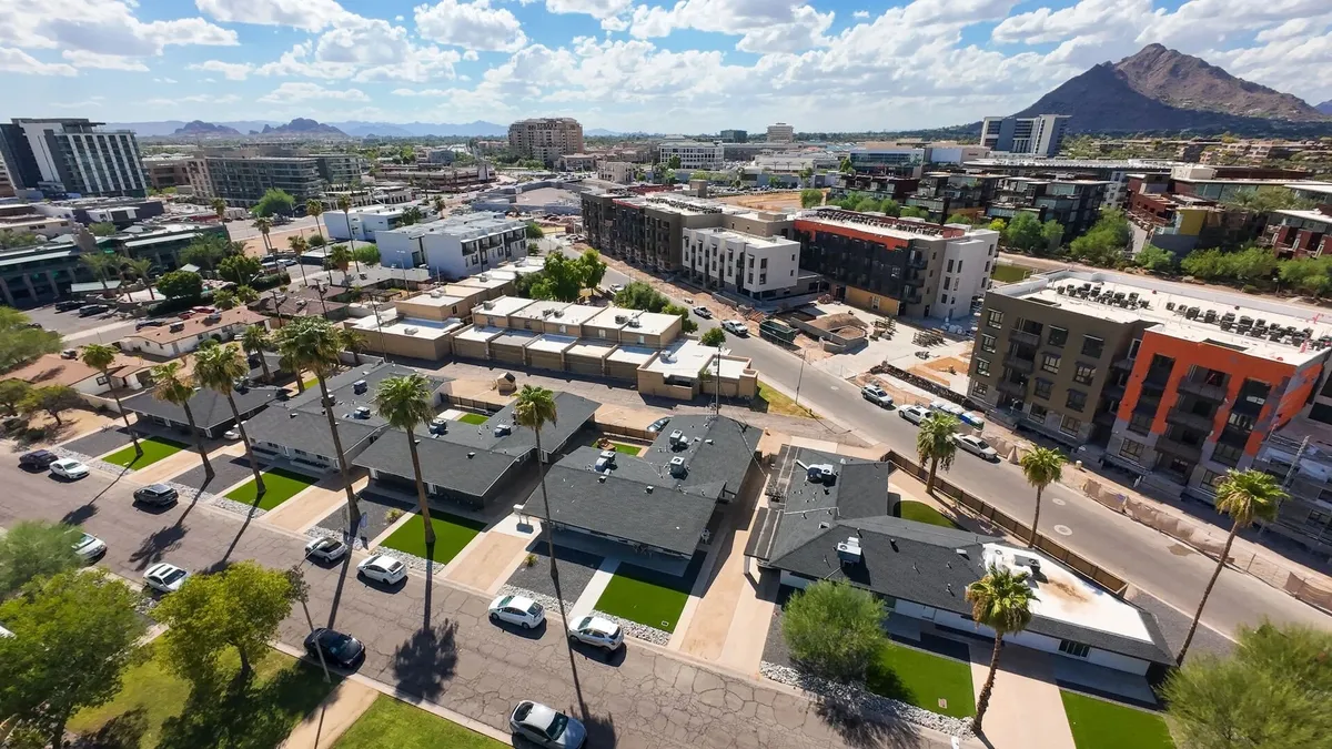A vibrant view of modern apartment buildings surrounded by green lawns and palm trees, with mountains in the distance and a lively parking area in the foreground. The scene feels active and urban, showing easy access to both natural beauty and city amenities.