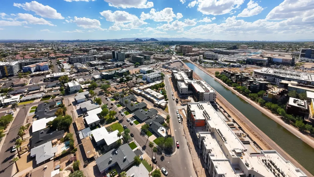 A sweeping urban landscape featuring a river, distant mountains, commercial buildings, and residential zones, with people walking along the river and exploring nearby shops and cafes. The scene is full of energy and activity, emphasizing Scottsdale’s dynamic city life.