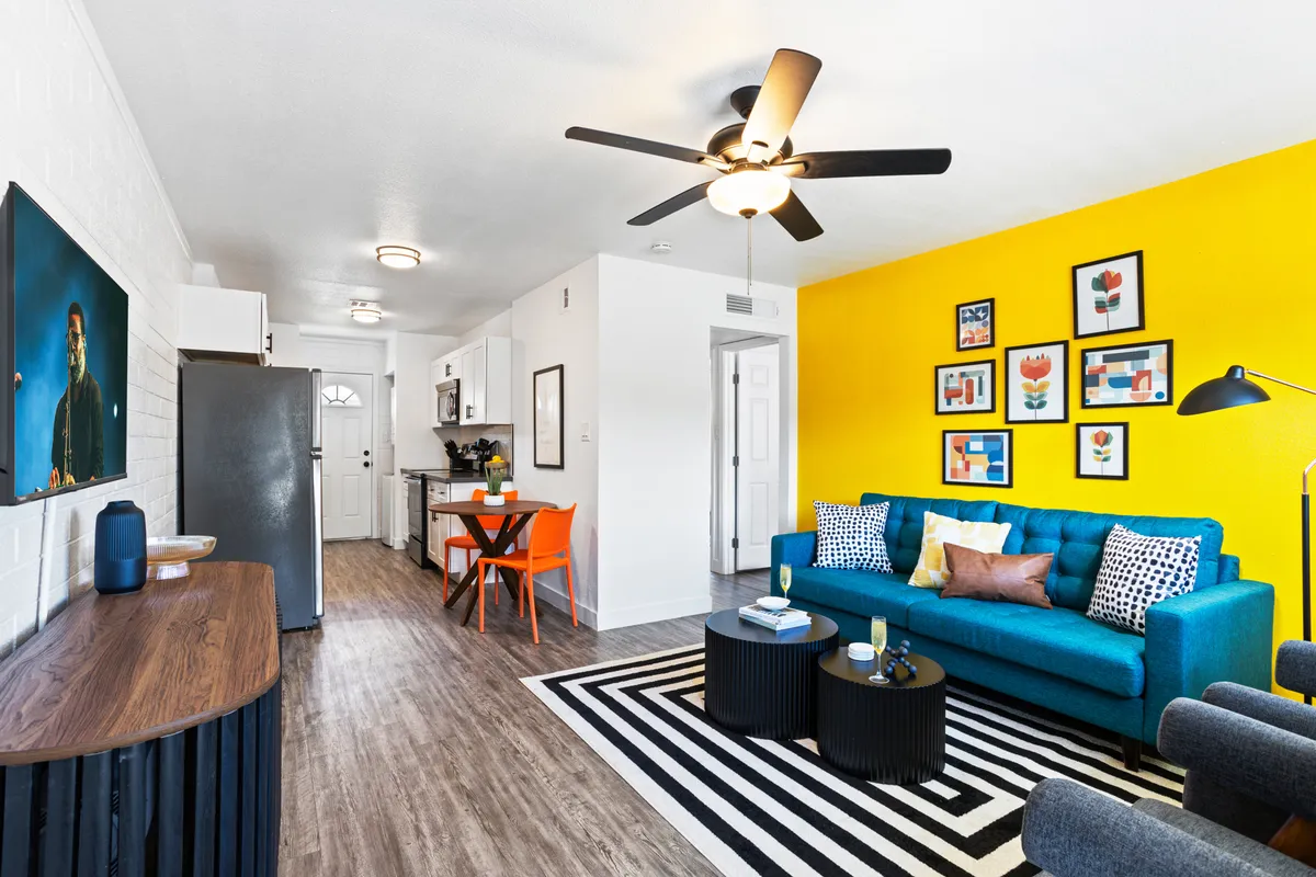 A bright and playful open-plan living and dining area featuring a yellow accent wall, a teal couch, a striped rug, colorful wall art, and a dining table with orange chairs. The space feels energetic and designed for both socializing and personal relaxation.